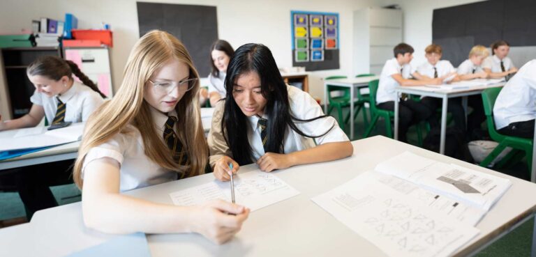 2 students sitting at a desk in a classroom. Both looking at a piece of paper on the desk.